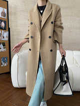 Person wearing a long beige coat holding a black handbag in an indoor setting.
