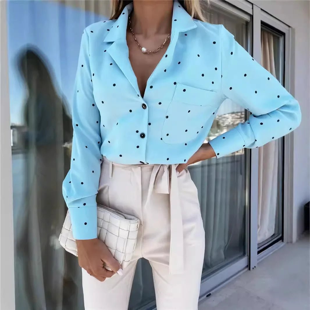 A woman wearing a light blue long-sleeve button-up blouse with black polka dots, paired with cream-colored high-waisted trousers and holding a white checkered clutch bag.