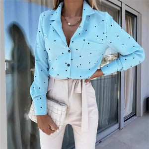 A woman wearing a light blue long-sleeve button-up blouse with black polka dots, paired with cream-colored high-waisted trousers and holding a white checkered clutch bag.